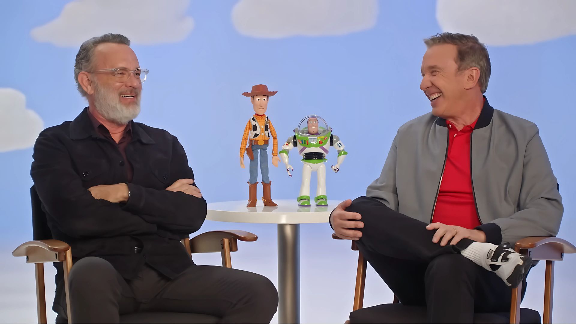 Tom Hanks and Tim Allen are sitting while Woody and Buzz toys are seen on a table in the middle