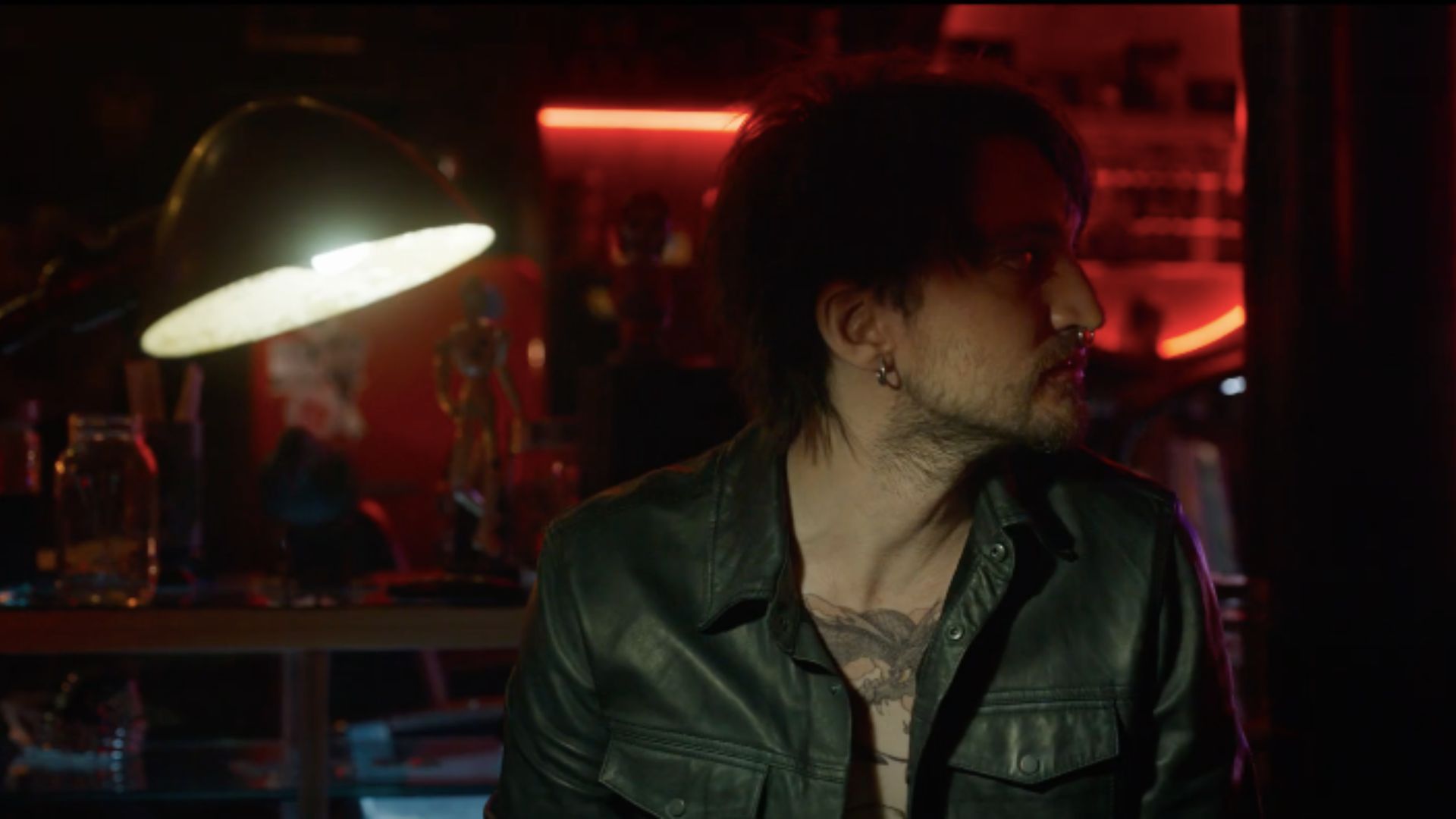 A still from Final Destination Bloodlines, with a tattooed man on the right, looking sideways and a lamp on his left. He is inside a tattoo parlour.