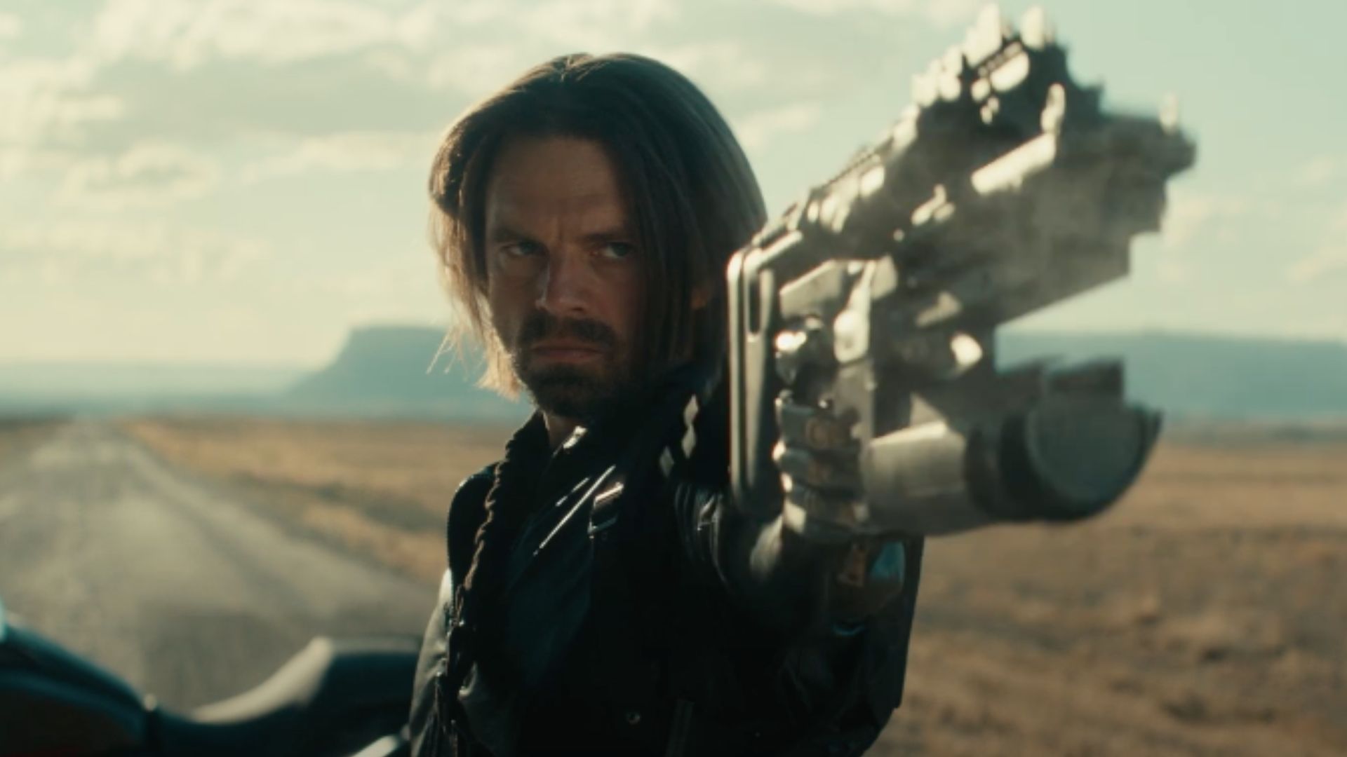 A still from Thunderbolts*, featuring Sebastian Stan as Bucky Barnes. It is a close shot of Barnes as he is pointing out a weapon to shoot. In the background there is a deserted road, and his bike.
