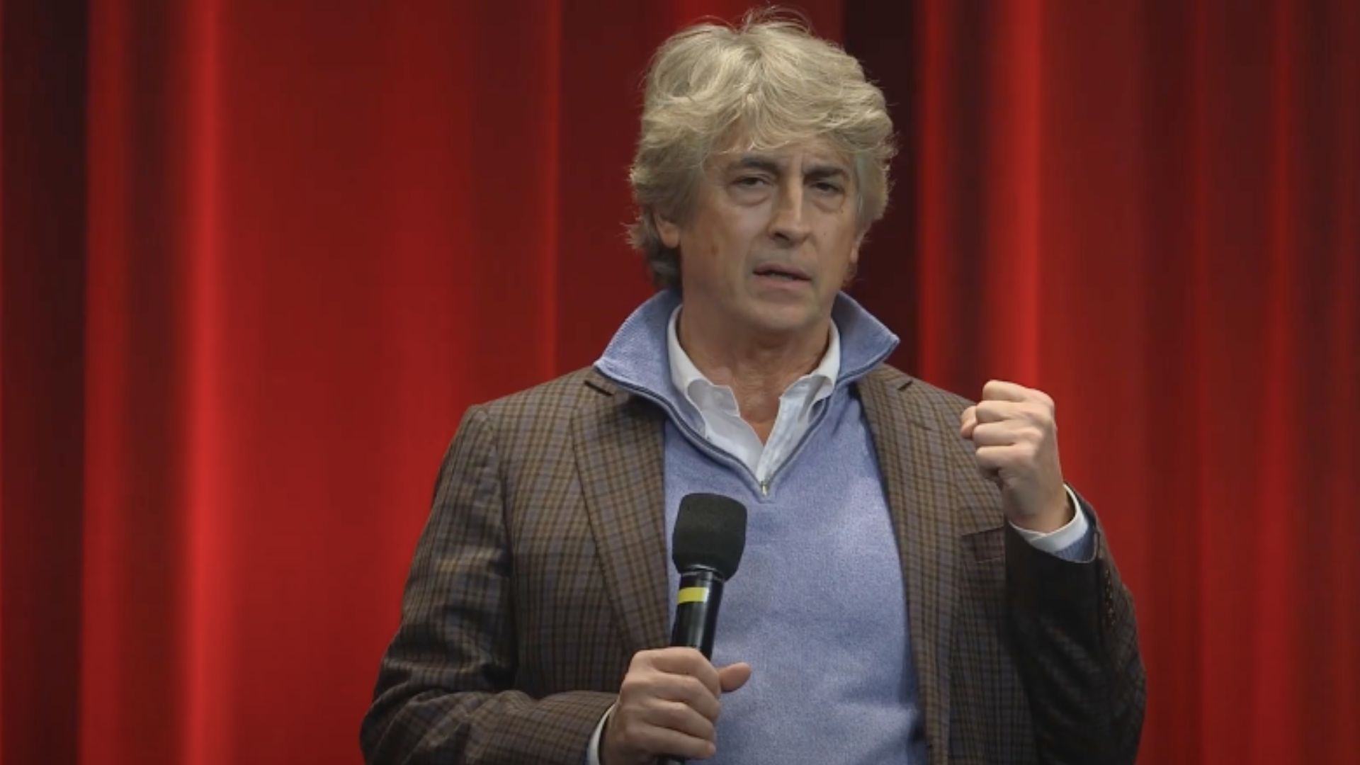 This is an image of filmmaker Alexander Payne. He is holding a mic on his right hand, and is holding a fist in his left. He is wearing a brown coat over powder blue shirt